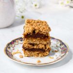 Two squares of date square stacked on a plate. Coffee mug a creamer cup in the background with white flowers.