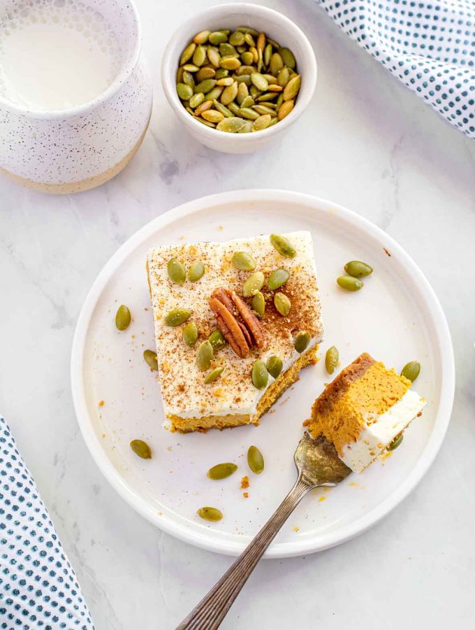 Shot from directly above of Square of pumpkin cheesecake, topped with whipped cream, a whole pecan and pumpkin seeds sitting on a white plate. A fork has cut a small piece off the corner and is also sitting on the plate. Small cup full of milk, and bowl full of pumpkin seeds in background.