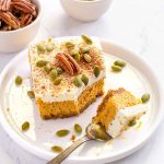 Square of pumpkin cheesecake, topped with whipped cream, a whole pecan and pumpkin seeds sitting on a white plate. Bowl of pecans and pumpkin seeds in background.