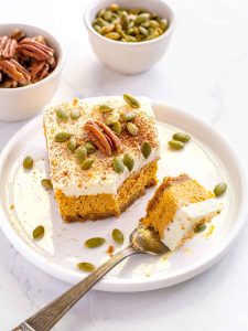 Square of pumpkin cheesecake, topped with whipped cream, a whole pecan and pumpkin seeds sitting on a white plate. Bowl of pecans and pumpkin seeds in background.