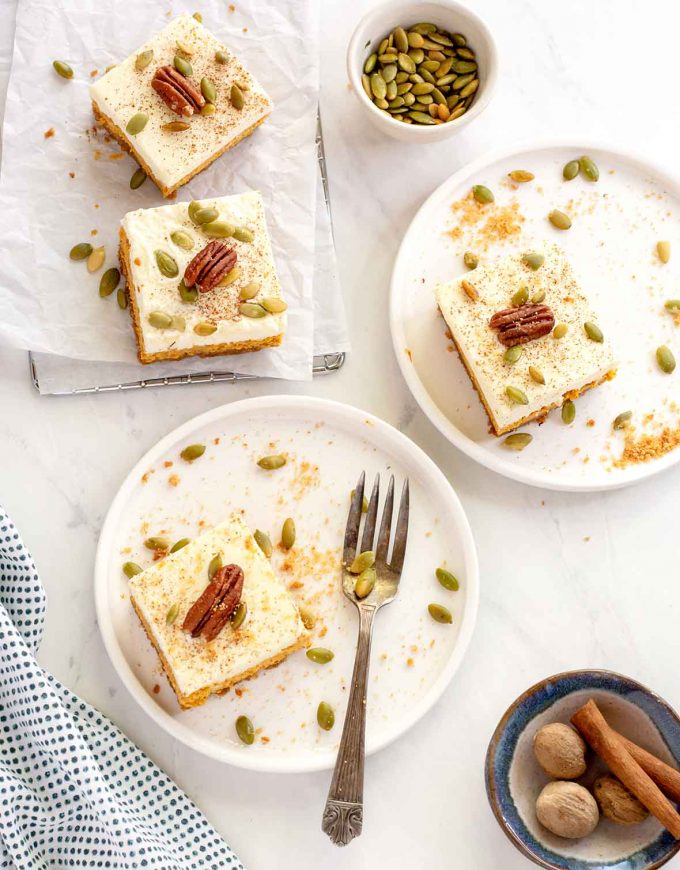 Shot from above, looking down at two white plates, each has a square of cheesecake on it. Pumpkin seeds are scattered on the plates. Top left corner has two other square on parchment paper, and a bowl of pumpkin seeds. 