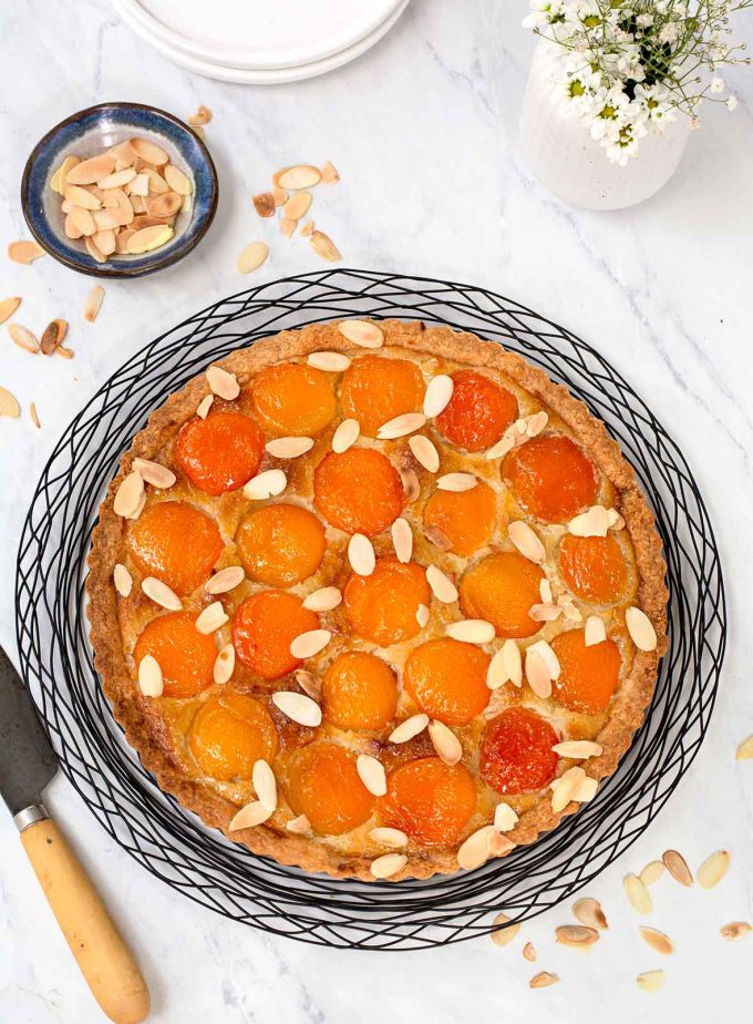 Round tart with apricot halves and sprinkled with sliced almonds. Tart is sitting on a round black wire rack. Small bowl of toasted almonds to the top left.