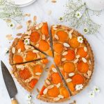 Round Tart with apricot halves in an almond filling, with sliced almonds scattered on top. Small white daisies scattered on top as well.