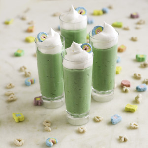 Four tall shot glasses filled with green pudding and topped with whipped cream and rainbows marshmallows