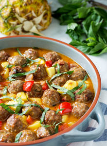 Round baking dish filled with meatballs, pineapple chunks, red peppers and chiffonade thai basil in a red curry sauce