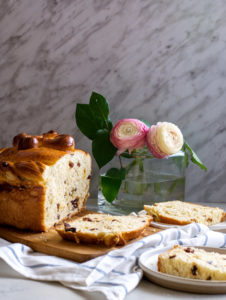 Large round loaf of bread with raisins in it with several large pieces cut out of it. Flowers in the background.