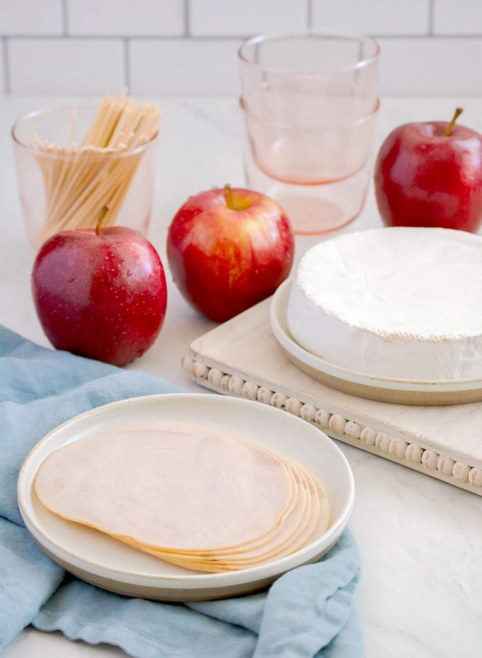 sliced deli turkey on a plate, three red apples and a round of brie cheese