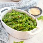 Small white serving dish filled with snap peas, sesame seeds in a small bowl in the background