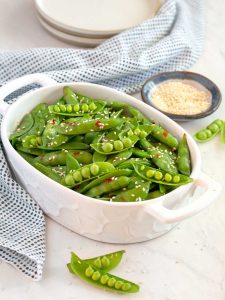 Small white serving dish filled with snap peas, sesame seeds in a small bowl in the background