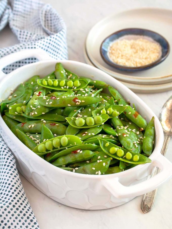 Small white serving dish filled with snap peas, with some pea pods open showing individual peas