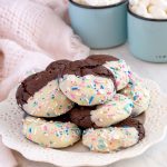 Small white plate with a tower of chocolate cookies on top. Each chocolate cookie is half dipped in white chocolate with sprinkles. Two cups of hot chocolate and marshmallows in background