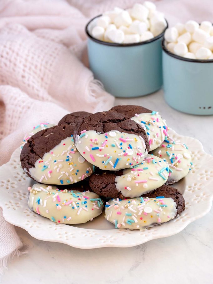 Small white plate with a tower of chocolate cookies on top. Each chocolate cookie is half dipped in white chocolate with sprinkles. Two cups of hot chocolate and marshmallows in background