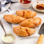 Four empanadas on a rectangle cooling rack, with two small dishes full of salsa and avocado sour cream