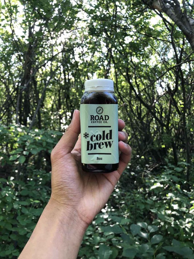 Small container of cold brew coffee held up in the air in a forest