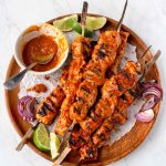 Cubes of cooked turkey on skewers, piled on a round wooden platter, with a small bowl of bbq sauce, limes and grilled red onions.