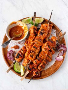 Cubes of cooked turkey on skewers, piled on a round wooden platter, with a small bowl of bbq sauce, limes and grilled red onions.