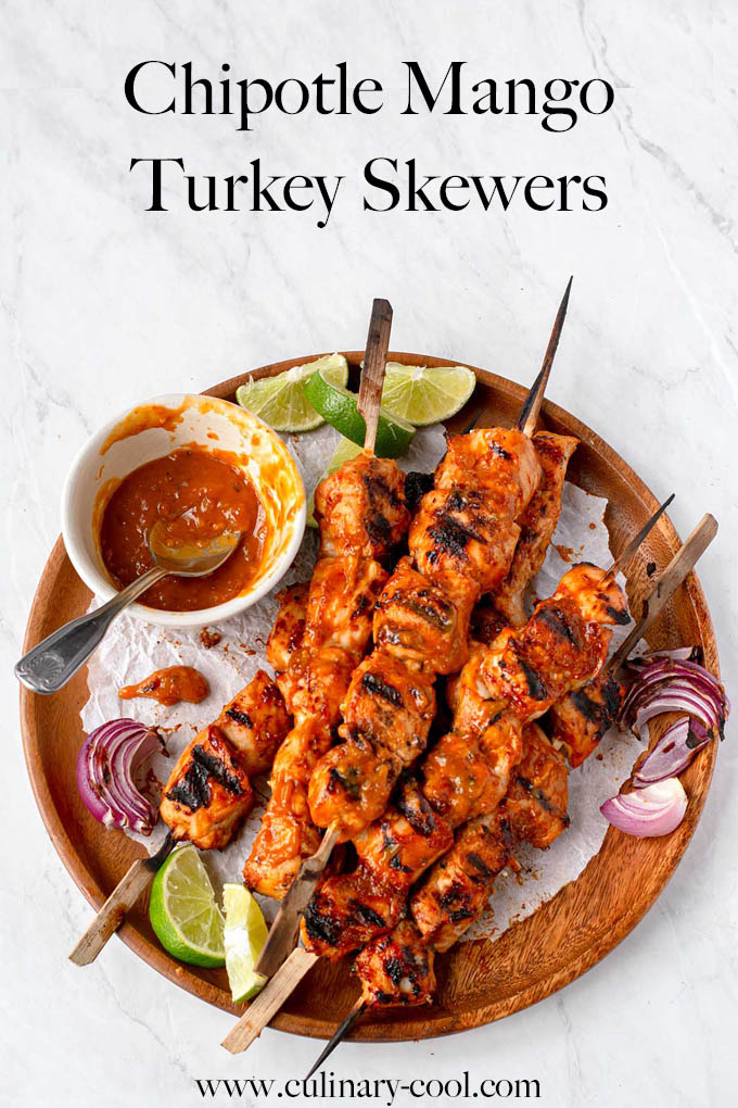 Cubes of cooked turkey on skewers, piled on a round wooden platter, with a small bowl of bbq sauce, limes and grilled red onions. Text at top of page that says "Chipotle Mango Turkey Skewers". Image for Pinterest