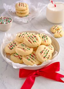 Round plate piled with shortbread cookies, topped with red, white and green sprinkles. There is a red bow in front of the plate. In the background is a small bowl full of sprinkles, and three cookies stacked next to a glass of milk.