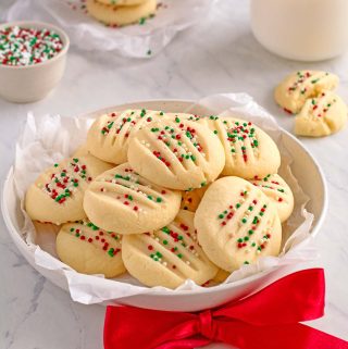 Round plate piled with shortbread cookies, topped with red, white and green sprinkles. There is a red bow in front of the plate. In the background is a small bowl full of sprinkles, and three cookies stacked next to a glass of milk.