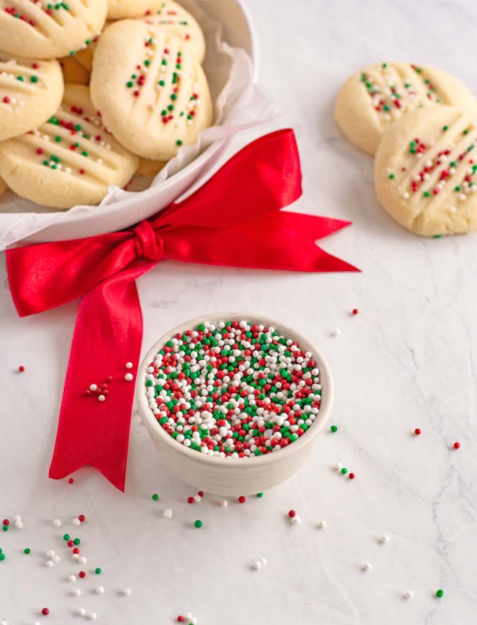 small bowl filled with red, white and green sprinkles. In the background is a plate piled high with shortbread cookies topped with the same sprinkles.