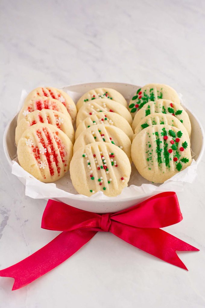 Round plate with three rows of shortbread cookies. First row has cookies with red sugar and white snowflake sprinkles. Second row is cookies with red, white and green sprinkles. Third row is cookies with green sugar and red and green mistletoe sprinkles.