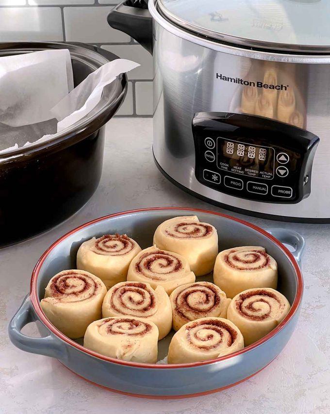 ten unbaked cinnamon rolls in a shallow pie dish. Slow cooker in the background.