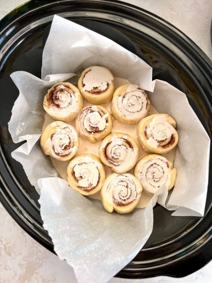 ten cinnamon rolls in an oval slow cooker covered lightly with cream.