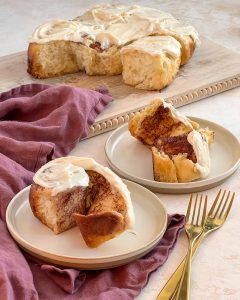 cinnamon roll covered with cream cheese icing sitting on a round plate. In the background the remaining 9 cinnamon buns sit on a cutting board.