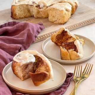 cinnamon roll covered with cream cheese icing sitting on a round plate. In the background the remaining 9 cinnamon buns sit on a cutting board.