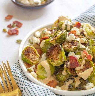 Small plate piled with roasted Brussels sprouts that were topped with pumpkin seeds, bacon pieces and crumbled feta. Drizzled with a tahini sauce.