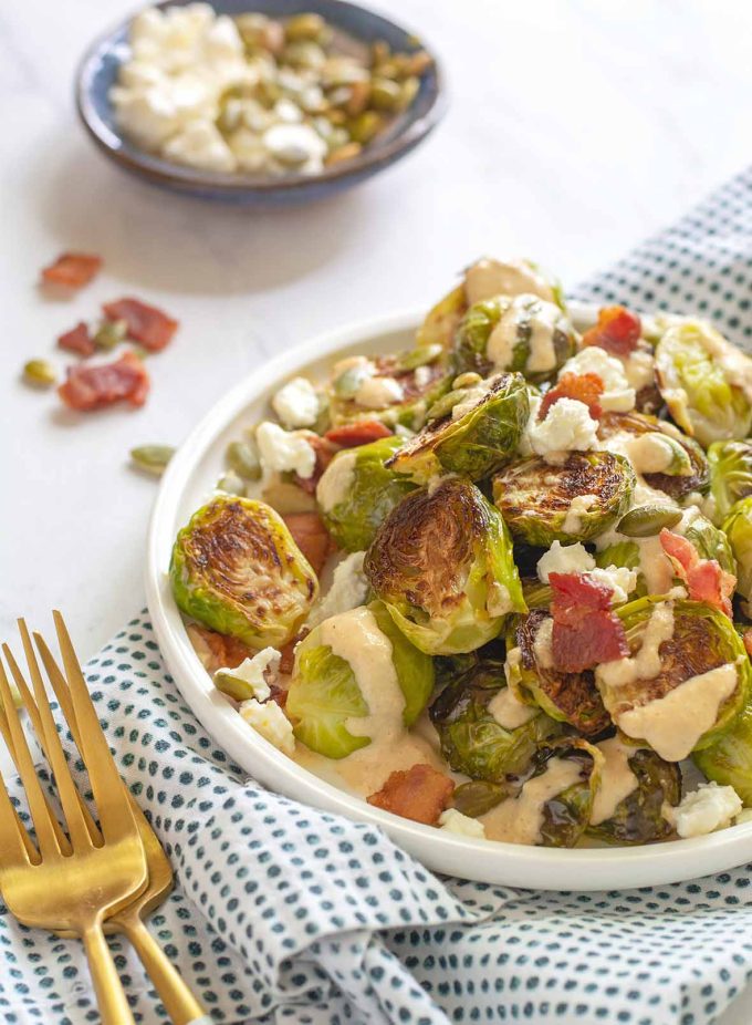 Small plate piled with roasted Brussels sprouts that were topped with pumpkin seeds, bacon pieces and crumbled feta. Drizzled with a tahini sauce.