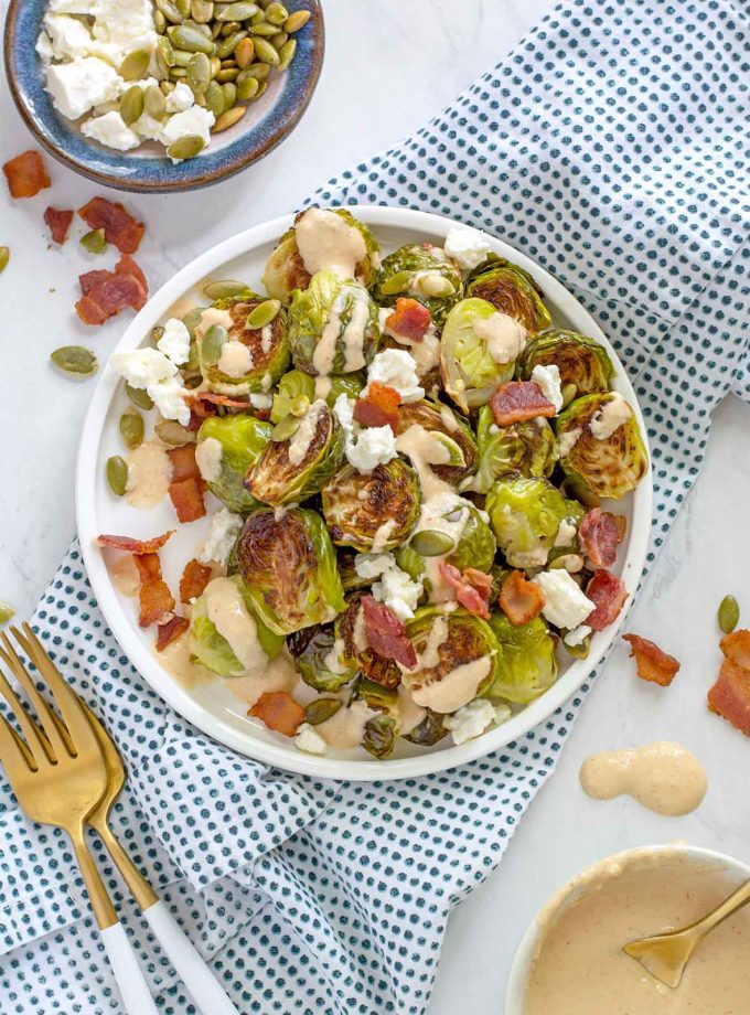 overhead shot of a plate full of halved Brussels sprouts that have been roasted until brown. Topped with bacon pieces, feta crumbles and drizzled with a tahini dressing.