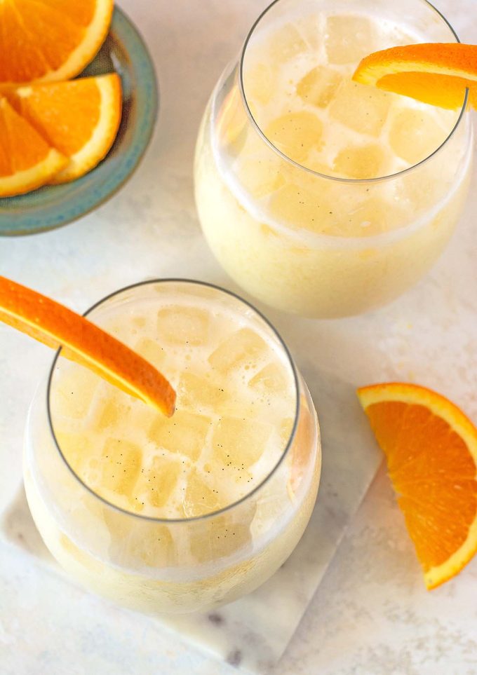 Top down shot of two cups filled with creamy orange creamsicle beverage. Orange slices on the rim of each cup. small blue bowl filled with sliced oranges in the corner.