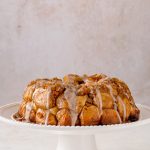 Baked Apple Fritter Monkey bread on a white cake stand with vanilla been glaze drizzled on top.