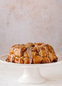 Baked Apple Fritter Monkey bread on a white cake stand with vanilla been glaze drizzled on top.