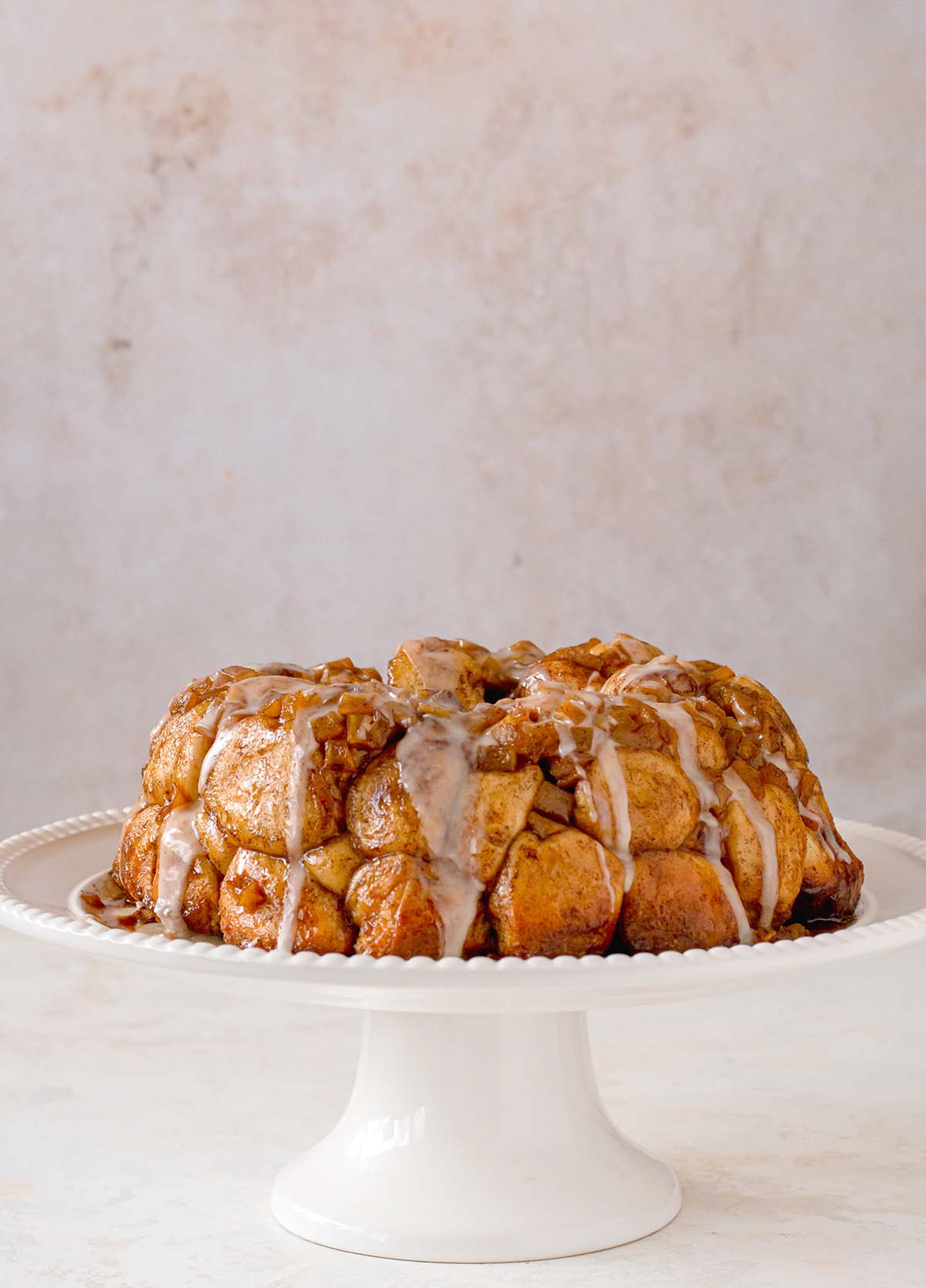 Baked Apple Fritter Monkey bread on a white cake stand with vanilla been glaze drizzled on top.
