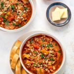 Top down shot of a bowl of cabbage rolls soup, sitting on a plate. Four slices of baguette nestled against the bowl. Small round dish in background with three pats of butter.