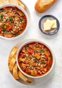 Top down shot of a bowl of cabbage rolls soup, sitting on a plate. Four slices of baguette nestled against the bowl. Small round dish in background with three pats of butter.