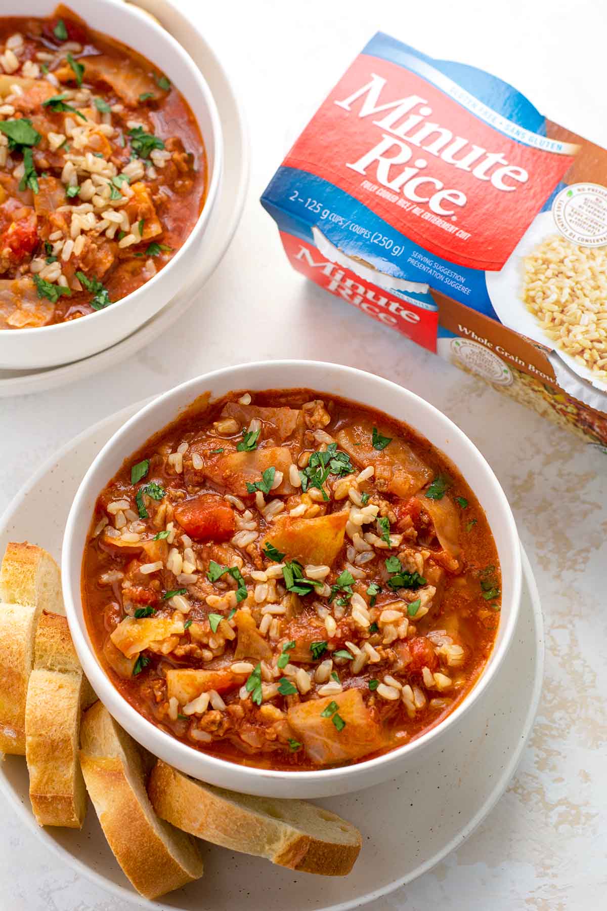 Bowl of cabbage roll soup on a plate with slices of baguette. Minute Rice single serve packaging in the top right corner.