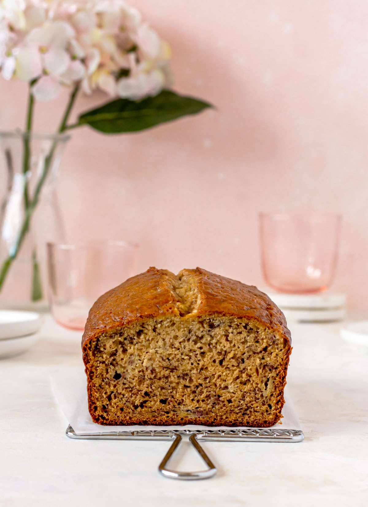 Loaf of banana bread sitting on parchment paper. Cups, flowers and plates in background.
